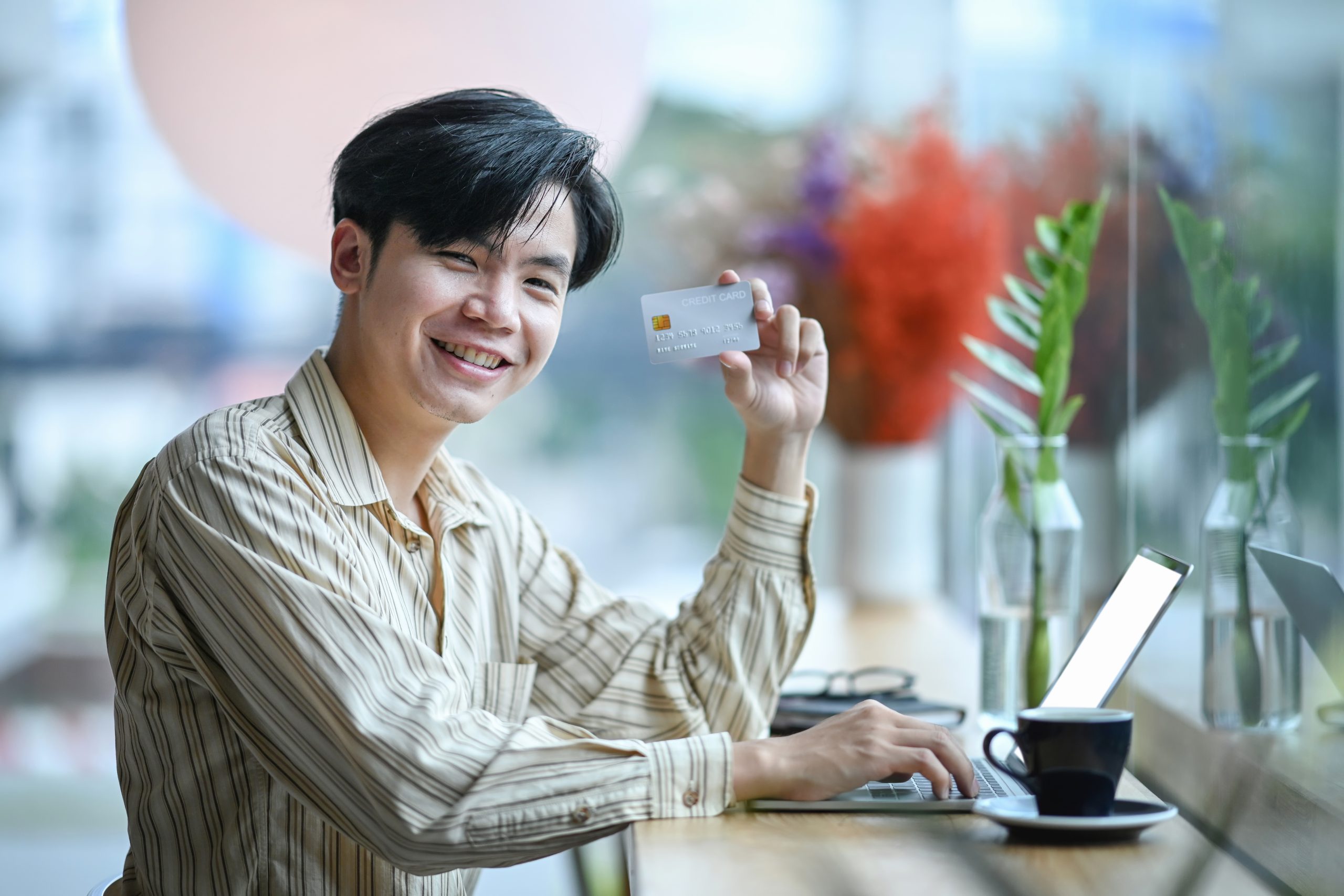 A man holding up a credit card in front of a laptop.
